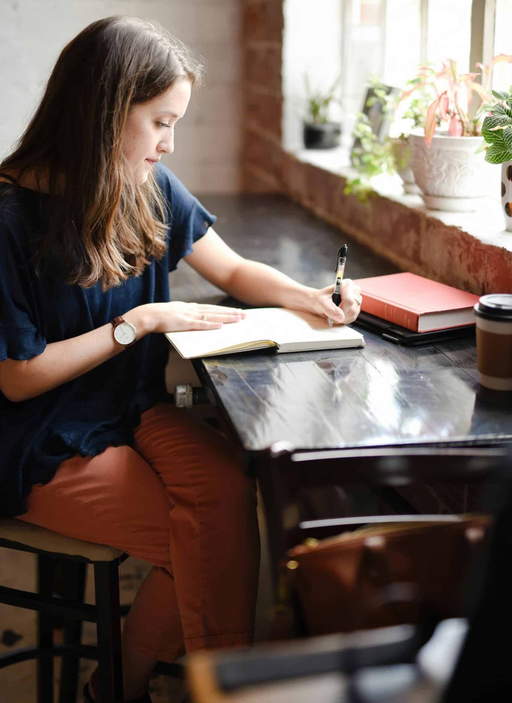 Young woman writing in a notebook at a desk by a window.